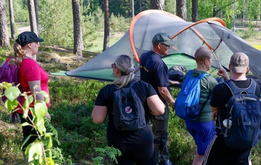 KUVA: Esko Soini / Keväinen mäntymetsä. EräKatri ja neljä retkeilijää tutustumassa tentsileen, mikä on viritetty puiden väliin..